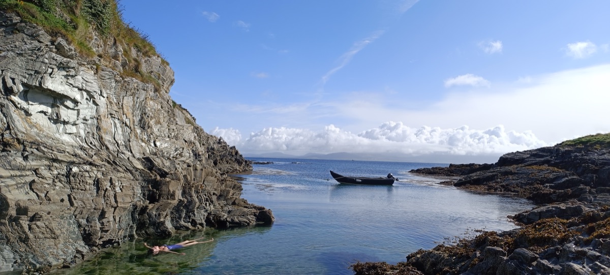 A swimmer floating in a sheltered rocky cove, currach moored nearby